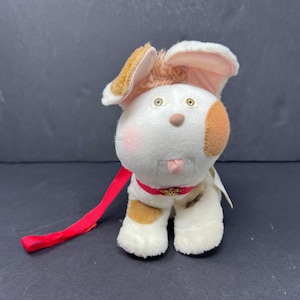 May include: A plush toy dog with white fur, brown spots, and floppy ears. It has a red collar and leash, with a pink tongue sticking out. The dog has a cute, friendly expression.