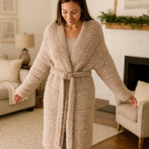 May include: A long, beige robe with a tie waist. The robe has a textured appearance and is worn over a light-colored top and gray pants. The person is wearing white sneakers. The background includes a living room with a fireplace and furniture.