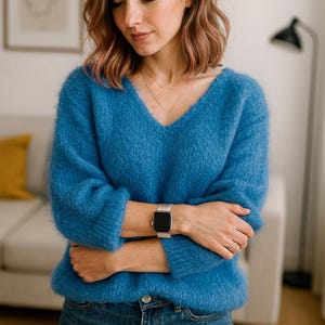 Könnte beinhalten: Eine Frau trägt einen leuchtend blauen, flauschigen V-Ausschnitt-Pullover und blaue Jeans. Eine silberne Smartwatch befindet sich am Handgelenk. Der Hintergrund umfasst ein weißes Sofa, ein gerahmtes Bild und eine Stehlampe.