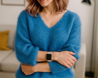 以下が含まれることがあります： 鮮やかなブルーのふわふわVネックセーターとブルージーンズを着た女性。手首にはシルバーのスマートウォッチ。背景には白いソファ、額入りの写真、フロアランプがあります。