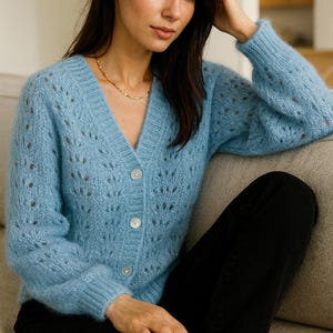 May include: A light blue cardigan with a V-neck and button closure. The cardigan has a delicate openwork design and is worn with a gold necklace and black pants. The model is sitting on a gray sofa.