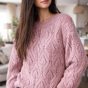 May include: A light pink knit sweater with a diamond pattern. The sweater has long sleeves and a crew neck. The model is wearing blue jeans. The sweater is made of a soft, textured yarn.