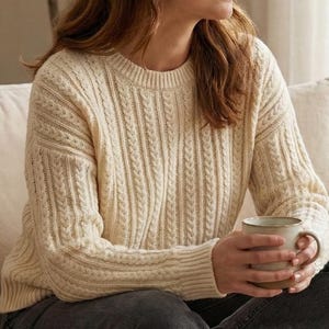 Op de afbeelding: Crèmekleurige kabelgebreide trui met een ronde hals en een textuurpatroon. De trui heeft lange mouwen. De persoon houdt een beige keramische mok vast. De persoon draagt een donkergrijze jeans.