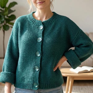 May include: A teal green, textured knit cardigan with a round neckline and large, matching buttons. The sleeves are three-quarter length with folded cuffs. The cardigan is paired with blue jeans. The background includes a living room setting with a plant and a coffee table.