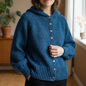May include: A blue hooded cardigan with a button-up front. The knitted sweater has a ribbed hem and cuffs, and silver-coloured buttons. The cardigan is a solid blue colour and has long sleeves.