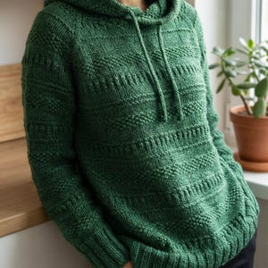 Puede incluir: Una sudadera con capucha verde tejida con cordón y un patrón texturizado. El suéter de manga larga tiene una capucha. La prenda está hecha de un material suave y cálido. Una prenda cómoda y elegante.