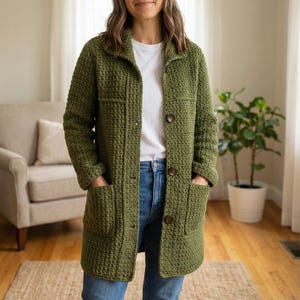 May include: A long, olive green crochet jacket with large brown buttons and two front pockets. The jacket has a collared neckline and is worn over a white t-shirt and blue jeans. The jacket is made of a textured, woven material.