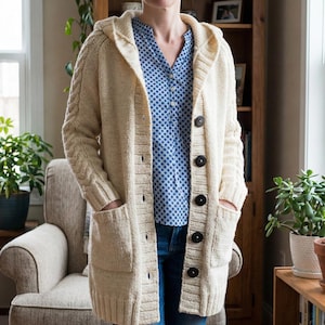May include: Cream-colored hooded cardigan with large black buttons and front pockets. The long-sleeved sweater features a cable knit design on the sleeves. The cardigan is buttoned up and worn over a blue patterned shirt and jeans.