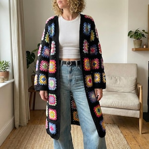 May include: A long, black crochet cardigan with colourful square patterns. The cardigan has an open front design and is made with a variety of bright colours, including pink, yellow, blue, and green. The garment is worn over a white top and blue jeans.