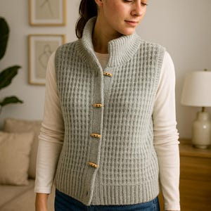 Pu&ograve; includere: Un gilet lavorato a maglia grigio chiaro con una trama a coste e un collo alto. Il gilet ha chiusure con bottoni in legno ed &egrave; indossato sopra una camicia bianca a maniche lunghe e jeans blu. Un capo elegante.
