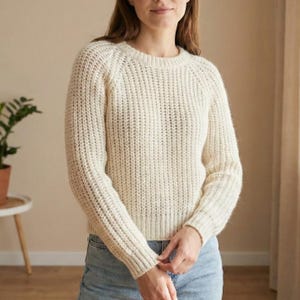 May include: Cream-colored, ribbed knit sweater with a crew neck and long sleeves. The sweater has a textured appearance and a relaxed fit. The model is wearing blue jeans. The sweater is a cozy, casual garment.
