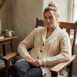 May include: Cream-colored cable-knit cardigan with a shawl collar and wooden buttons. The long-sleeved sweater features a textured pattern and is worn by a person seated in a wooden rocking chair. The person is wearing dark jeans.