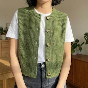 Op de afbeelding: Een groene gebreide vest met een ronde hals en een knoopsluiting. Het vest is effen groen van kleur en heeft meerdere bruine knopen. Het vest wordt over een wit t-shirt gedragen.