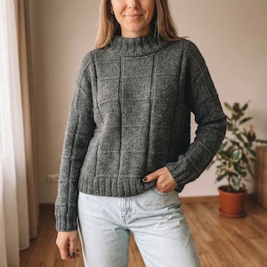 Op de afbeelding: Een grijze gebreide trui met een hoge halslijn en een textuurpatroon in rastervorm. De trui heeft lange mouwen en een geribbelde zoom. Het model draagt lichtblauwe jeans. De trui is gemaakt van een zacht, warm materiaal.