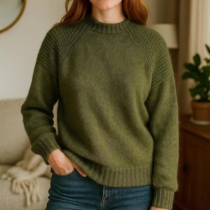 May include: An olive green sweater with a ribbed crew neck and cuffs. The long-sleeved sweater has a textured knit design on the shoulders and sleeves. The person in the image is wearing blue jeans.