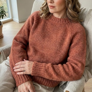 Peut inclure: Un pull de couleur rouille avec un col rond et des manches longues. Le tricot semble doux et confortable, avec des poignets côtelés et une coupe décontractée. Le pull est porté par une personne assise dans une chaise.