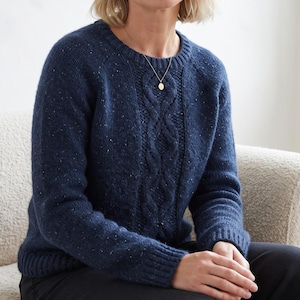May include: A navy blue long-sleeved jumper featuring a speckled texture and a central vertical cable knit pattern. The garment has a round ribbed neckline and ribbed cuffs, crafted from a knit fabric with subtle flecks of lighter blue and white.