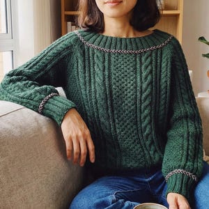 May include: A forest green cable-knit sweater with a crew neck and long sleeves. The sweater features a decorative trim around the neckline and cuffs. The person is wearing blue jeans. The sweater is a cozy, handmade garment.