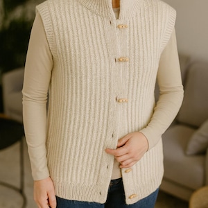 Pu&ograve; includere: Gilet in maglia a coste color crema con collo alto e chiusure a toggle in legno. Il maglione senza maniche &egrave; indossato sopra una camicia beige a maniche lunghe e jeans blu. Il gilet ha un motivo a coste verticali.