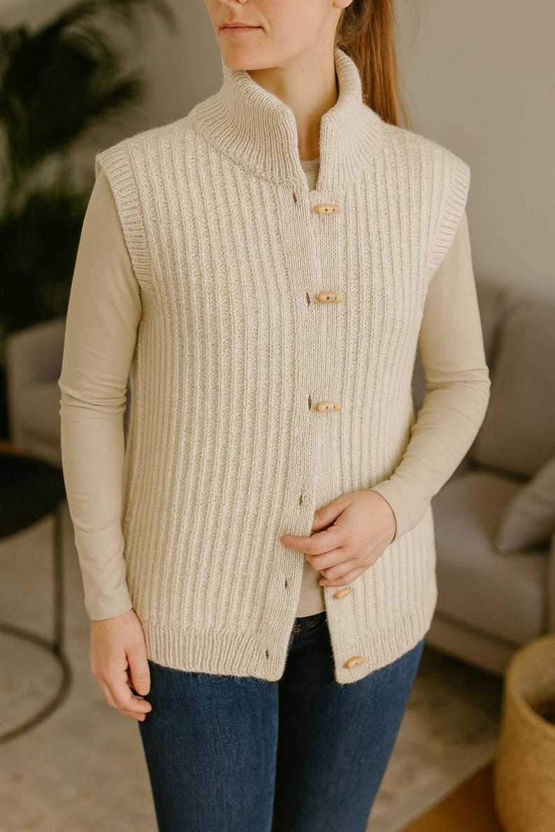 Pu&ograve; includere: Gilet in maglia a coste color crema con collo alto e chiusure a toggle in legno. Il maglione senza maniche &egrave; indossato sopra una camicia beige a maniche lunghe e jeans blu. Il gilet ha un motivo a coste verticali.