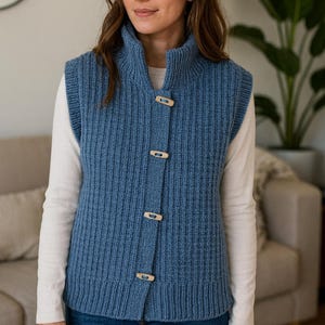 Pu&ograve; includere: Un gilet blu lavorato a maglia con collo alto e chiusure a toggle in legno. L'indumento senza maniche &egrave; indossato sopra una camicia bianca a maniche lunghe e abbinato a jeans blu. Il gilet ha una trama a coste.