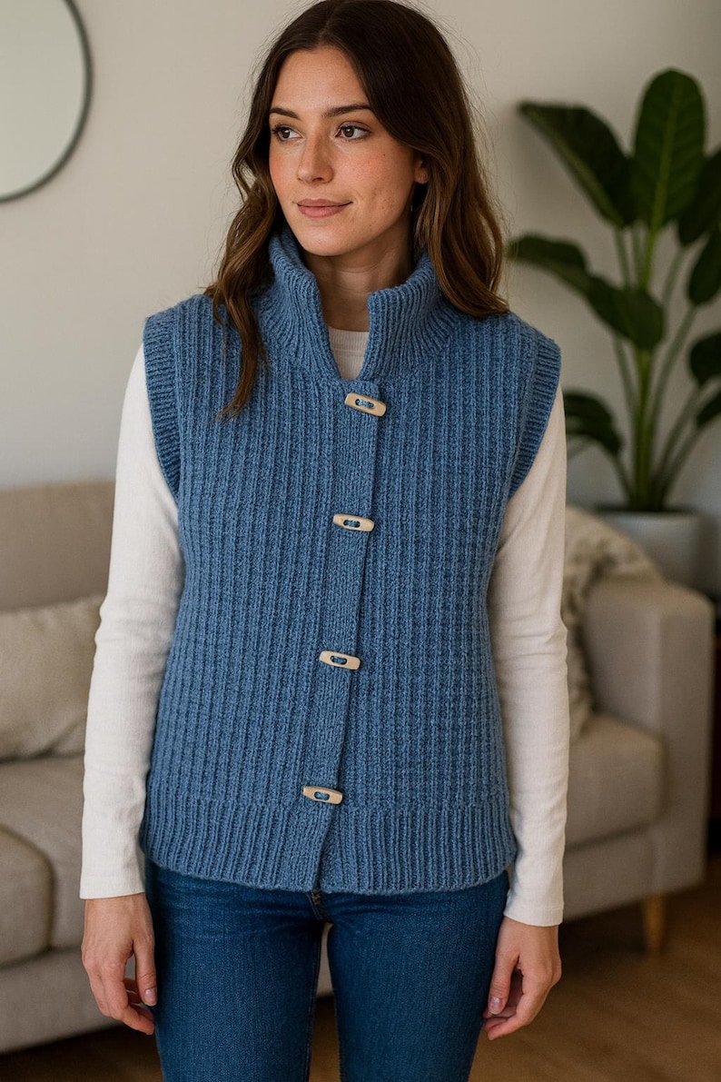Pu&ograve; includere: Un gilet blu lavorato a maglia con collo alto e chiusure a toggle in legno. L'indumento senza maniche &egrave; indossato sopra una camicia bianca a maniche lunghe e abbinato a jeans blu. Il gilet ha una trama a coste.