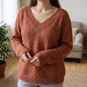 Puede incluir: Un suéter color óxido con cuello en V profundo. El suéter de manga larga tiene un diseño de punto texturizado y un dobladillo y puños acanalados. Las mangas presentan un detalle de punto trenzado. El suéter está hecho de un material suave y cálido.