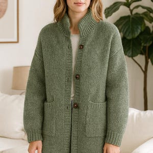 Puede incluir: Un cárdigan de punto verde con cuello alto y bolsillos delanteros grandes. El cárdigan tiene un cierre de botones con tres botones marrones. Las mangas son largas y el material parece ser un tejido grueso y cálido.