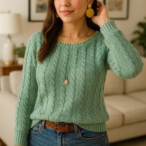 Op de afbeelding: Een lichtgroene kabelgebreide trui met een gele halslijn, gecombineerd met een blauwe jeans en een bruine riem. Het model draagt gele oorbellen en een hangerketting. De trui heeft lange mouwen en een klassiek ontwerp.