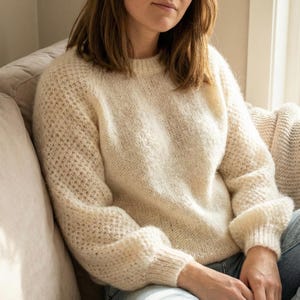 May include: A cream-coloured knitted jumper with a round neckline and long sleeves. The sleeves have a textured pattern and ribbed cuffs. The jumper appears soft and comfortable. The model is wearing blue jeans.