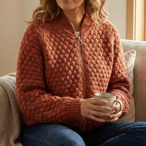May include: A rust-colored, textured knit bomber jacket with a full zipper closure. The jacket features a diamond pattern and ribbed collar and cuffs. The person is holding a beige mug. The jacket is worn with blue jeans.