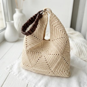 Może przedstawiać: Kremowa torba typu tote wykonana na szydełku z geometrycznym wzorem. Torba posiada brązową, marszczoną skórzaną rączkę przymocowaną srebrnym pierścieniem. Torba leży na białej powierzchni.