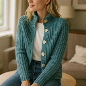 May include: A teal ribbed knit cardigan with a high collar and large, round, off-white buttons. The cardigan is worn over a white t-shirt and paired with blue jeans. The person is sitting on a wooden stool.