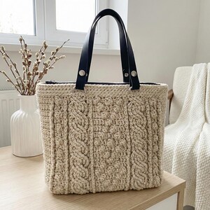 Puede incluir: Un bolso tote beige tejido a ganchillo con asas de cuero negro. El bolso presenta un diseño de punto trenzado texturizado y está asegurado con herrajes metálicos plateados. El bolso está sobre una superficie de madera clara.