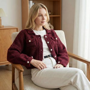 Puede incluir: Chaqueta de punto texturizado color burdeos con cuello y botones perlados. La chaqueta tiene dos bolsillos delanteros y se usa con una camiseta blanca y pantalones claros. Se ve un cinturón negro.