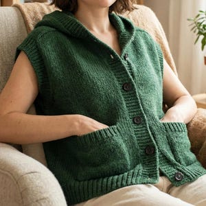 May include: A forest green hooded vest with large, dark brown buttons and two front pockets. The sleeveless sweater is made of a thick knit material. The vest is buttoned up and has a hood.