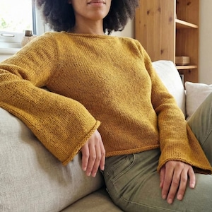 May include: Mustard yellow knitted sweater with flared sleeves. The sweater features a boat neck and a ribbed hem. The sleeves are long and flared at the wrist. The person is wearing olive green trousers.