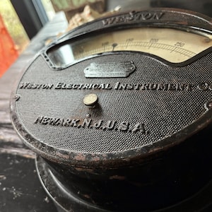 Puede incluir: Primer plano de un medidor vintage de Weston Electrical Instrument Co. El medidor tiene una carcasa de metal oscura y texturizada con el nombre de la empresa y "Newark, N.J. U.S.A." en relieve. La esfera del medidor muestra números hasta 250.