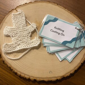 May include: A white knitted teddy bear on a wooden surface with a card that says "Knitting Casting On".