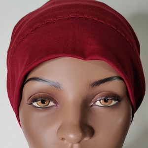 May include: A deep burgundy beanie cap made of a soft, smooth fabric. The beanie features a simple, fitted design that covers the crown of the head, with a visible horizontal seam detail running across the front band of the hat, providing a clean look.