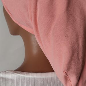 May include: A light pink head covering, possibly a bonnet or cap, made of soft fabric. The head covering is worn over a white ribbed shirt. The image is a close-up of the back of a person.