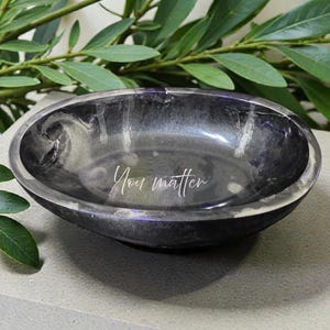 May include: A dark gray, oval-shaped decorative bowl with a marbled effect. The words "You matter" are inscribed in white on the inside of the bowl. The bowl is set on a light gray surface, with green leaves in the background.