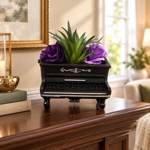 May include: A black piano-shaped planter featuring a decorative silver filigree accent on the front panel. The planter holds a green succulent plant flanked by two large, vibrant purple artificial flowers, all resting on four small, rounded feet.