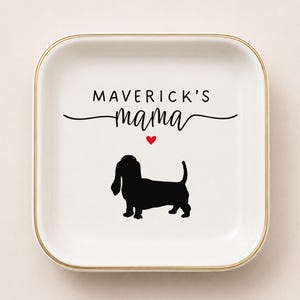 Puede incluir: Un plato cuadrado de cerámica blanca con un borde dorado. El centro presenta el texto negro "MAVERICK'S mama" sobre un pequeño corazón rojo y la silueta negra de un perro salchicha. El diseño está centrado en la superficie blanca lisa.