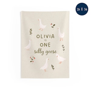 May include: A beige wall hanging with the text "Olivia is One silly goose" and illustrations of white geese, leaves, and flowers. The wall hanging measures 26 x 36 inches.