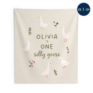 May include: A beige tapestry with the text "OLIVIA IS ONE silly goose" surrounded by illustrations of white geese, sprigs, and berries. The tapestry measures 88 x 104 inches.