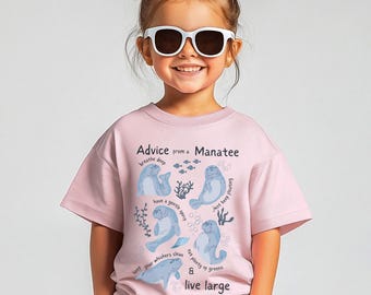 Peut inclure: Un t-shirt rose clair avec le texte "Advice from a Manatee" et des illustrations de lamantins. Le t-shirt comporte également des phrases comme "breathe deep" et "live large". L'enfant porte des lunettes de soleil blanches.