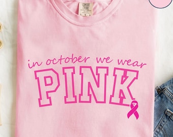 May include: A light pink t-shirt with the text "in october we wear PINK" in a bold, pink font. A pink ribbon graphic is included. The shirt is folded, with a portion of blue jeans visible in the background.