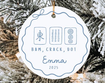 Puede incluir: Adorno navideño redondo de cerámica blanca con borde festoneado azul y cuerda dorada. Presenta un diseño de fichas de Mahjong, el texto "BAM, CRACK, DOT", el nombre "Emma" y el año "2025".