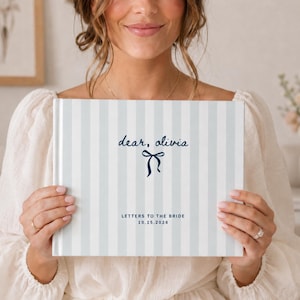 May include: A hardcover book with a light blue and white vertical striped cover. The front features the dark blue text "dear, olivia" above a dark blue bow illustration, followed by "LETTERS TO THE BRIDE" and the date "10.15.2026".